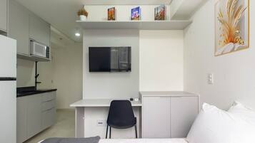 Quarto compacto com cama, escrivaninha e cadeira, televisão, micro-ondas e um pequeno armário.