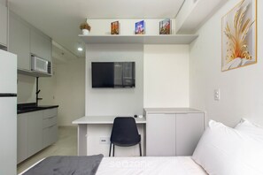 Quarto compacto com cama, escrivaninha e cadeira, televisão, micro-ondas e um pequeno armário.
