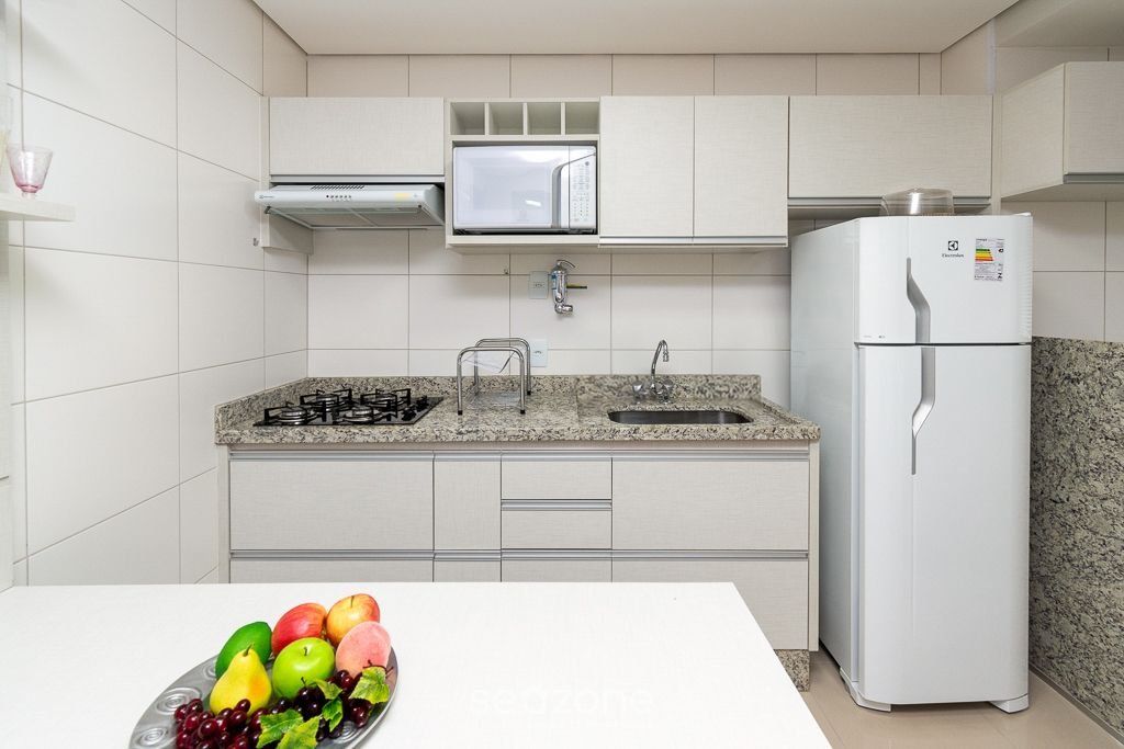Uma cozinha moderna com armários brancos, bancada de granito, micro-ondas embutido, fogão a gás e geladeira branca.