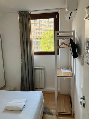 Habitación de hotel con cama, radiador, ventana con cortinas, televisor montado en la pared y estante de madera.