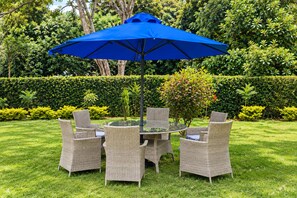 A round glass table with wicker chairs and a blue umbrella in a well-maintained garden. - Royal Gardens Muthatari - Embu : Standard Double Room with Two Double Beds (Embu)