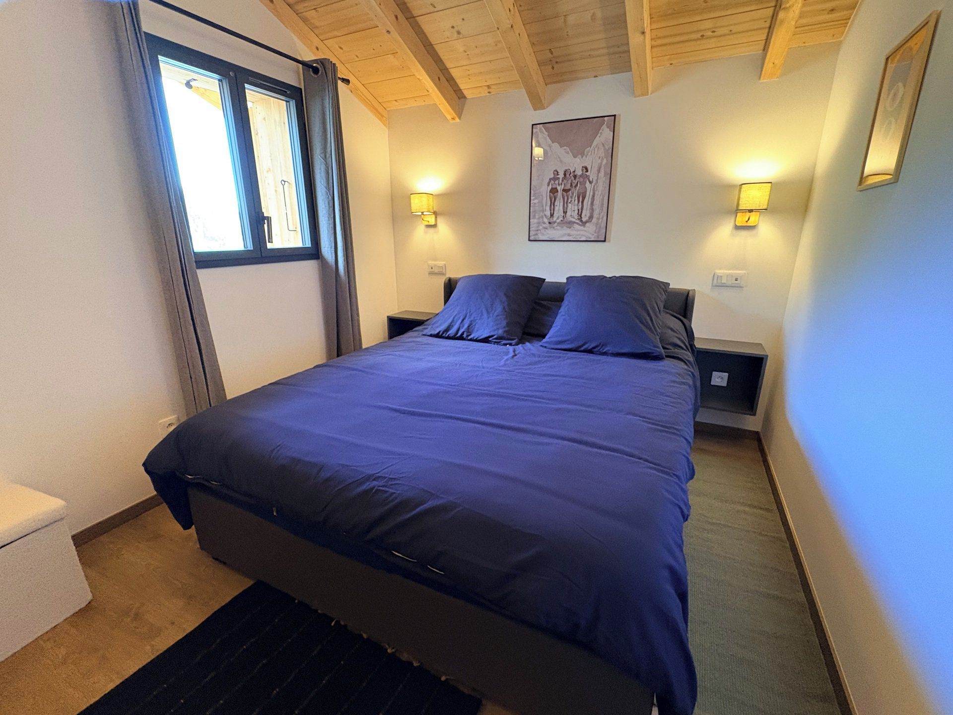 A bedroom with a large bed, two bedside tables, a window with curtains, and a wall-mounted picture.