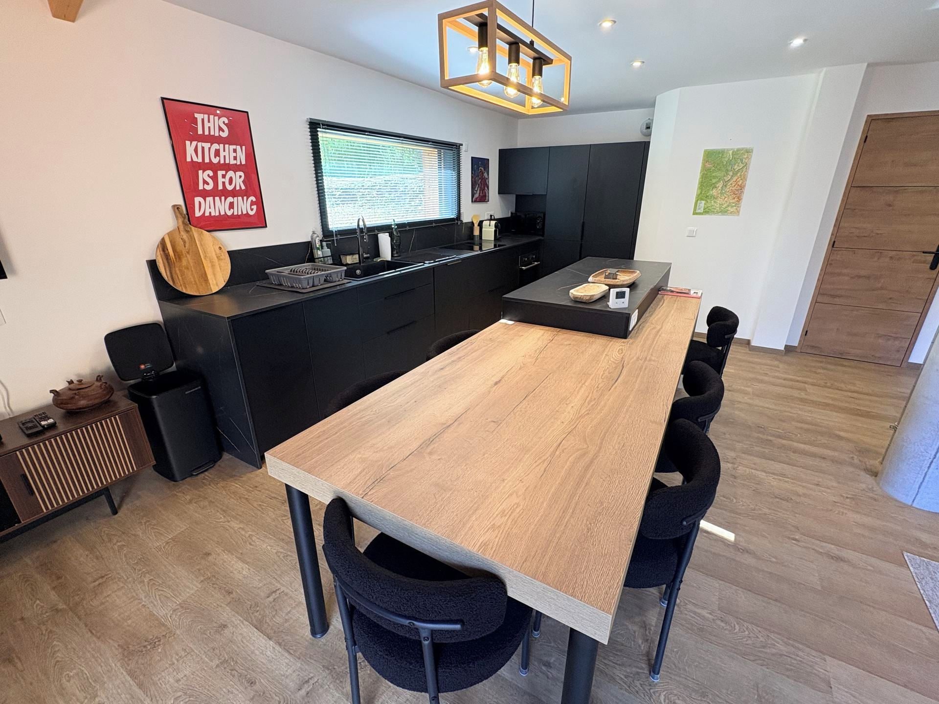 Une cuisine moderne dotée d’un îlot central, de meubles de rangement noirs et d’un coin repas avec une longue table en bois et des chaises noires.