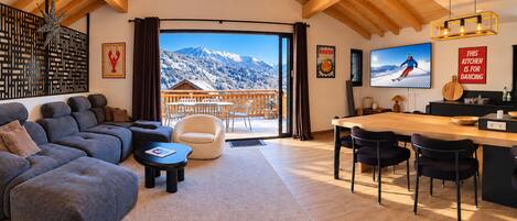 Un salon moderne avec un grand canapé, une table basse et un coin repas avec des chaises. Une grande fenêtre offre une vue sur les montagnes.