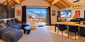 A modern living room with a large sofa, a coffee table, and a dining area with chairs. There is a view of mountains through the large window. - Chalet Cap'Sud - Two Bedroom Chalet, Sleeps 4 (Vaujany)