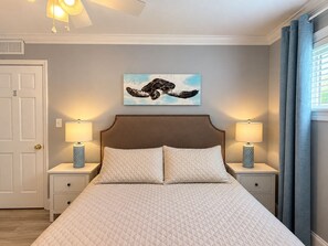 A bedroom with a bed, two bedside tables, a wall-mounted artwork, and a window with curtains. - Surf and Sand Retreat-Walk to Beach, Modern Amenities (Virginia Beach)