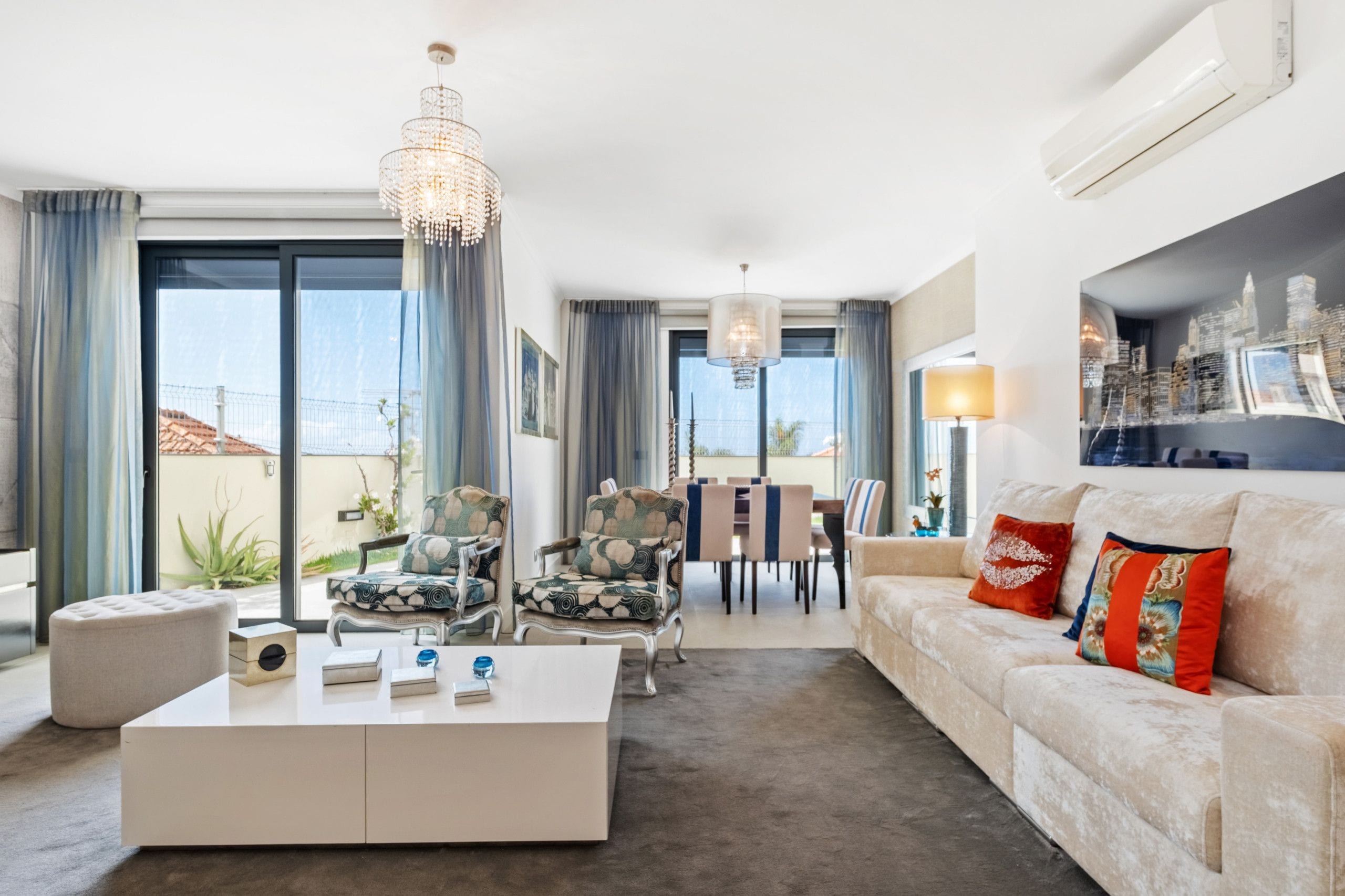A modern living room with a white sofa, a coffee table, and a chandelier. There is a dining area with chairs and a table.