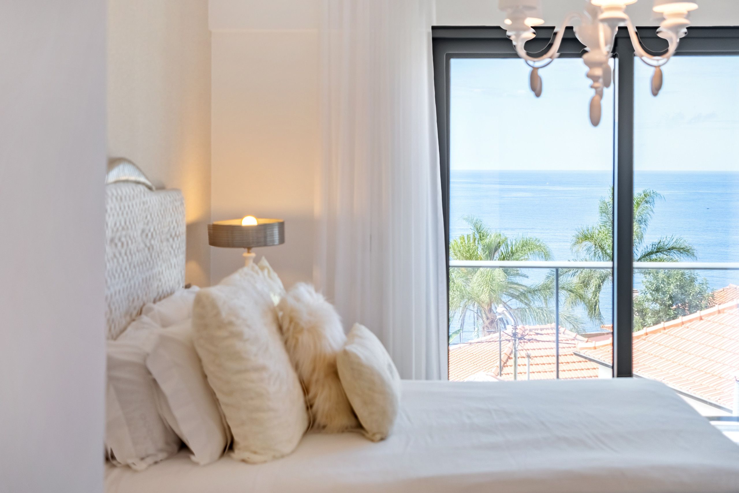 A bedroom with a large bed, a nightstand with a lamp, and a view of the ocean through a large window.