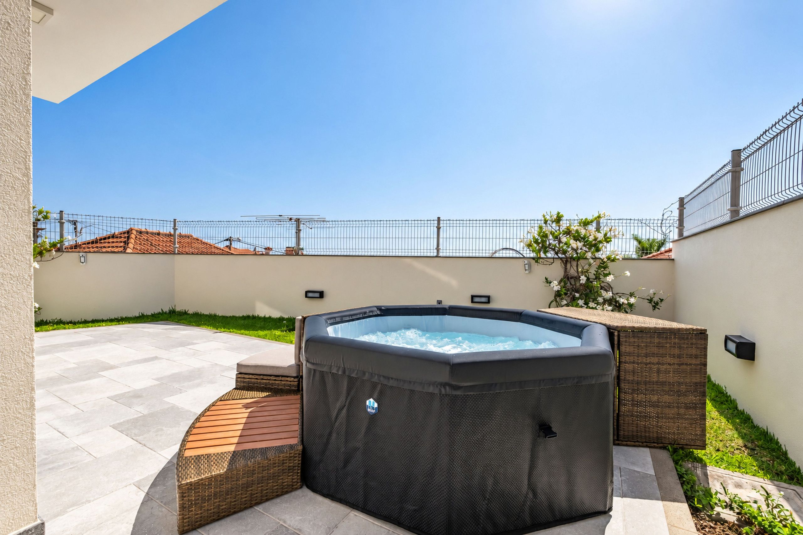 Een jacuzzi in een terras buiten, met een heldere lucht, een metalen hek en een gebouw met pannendak op de achtergrond.