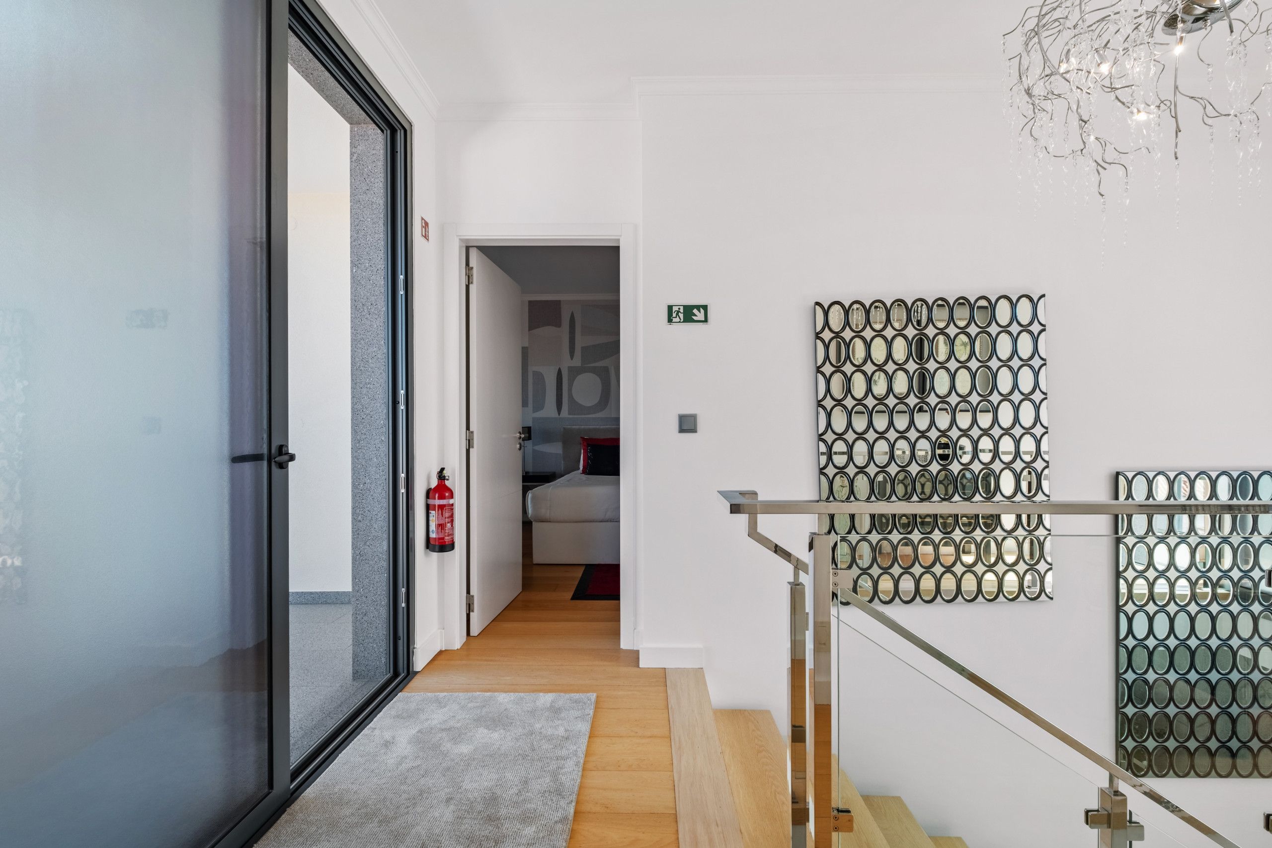 Un couloir d’hôtel avec une porte en verre, un extincteur, un escalier doté d’une rampe en métal et un mur orné d’un motif de miroirs.