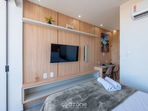 Ein modernes Hotelzimmer mit einem an einer Holzwand montierten Flachbildfernseher, einer kleinen Pflanze auf einem Regal, einem Gemälde und einem ordentlich bezogenen Bett mit einem zusammengerollten Handtuch.