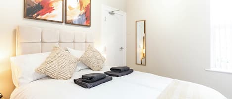 Een hotelkamer met een groot bed, twee kussens, een opgevouwen handdoek, een spiegel en twee abstracte schilderijen aan de muur.