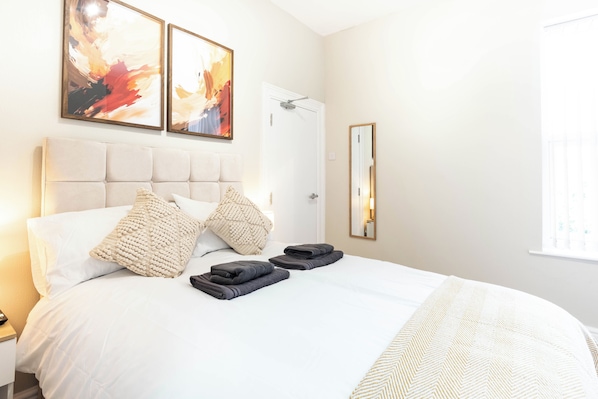 Ett hotellrum med en stor säng, två kuddar, en hopvikt handduk, en spegel och två abstrakta målningar på väggen.