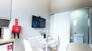 Ein modernes Hotelzimmer mit einem an der Wand befestigten Flachbildfernseher, einem kleinen runden Tisch mit einem weißen Stuhl und einer Duschkabine aus Glas.