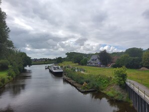 A river with a boat docked on the right, a house with a porch on the left, and a grassy area with trees in the background.