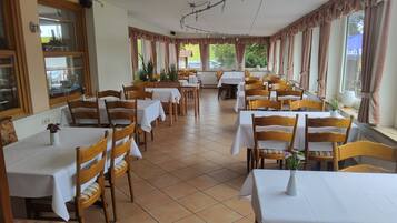 En restaurant med flere borde og stole, store vinduer med gardiner, og et flisegulv.