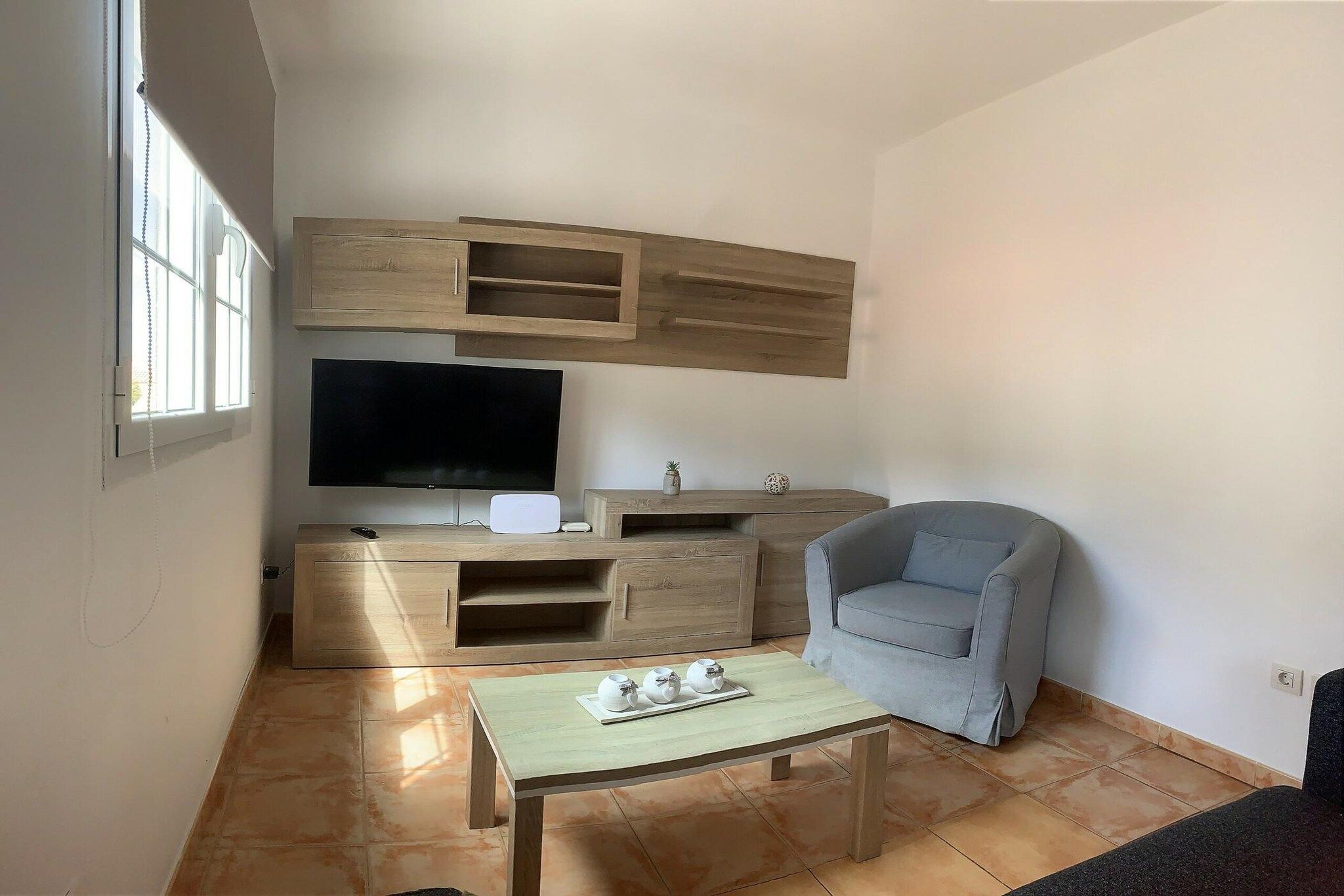 A living room with a TV, wooden entertainment unit, a grey armchair, and a small table with cups.