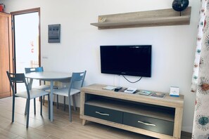 Ein Zimmer mit Esstisch, Stühlen, einem an der Wand befestigten Fernseher, einer Holzreolen über dem Fernseher und einer kleinen Kommode mit Schubladen.