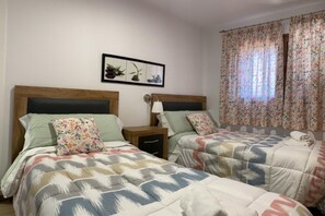 Une chambre double avec deux lits, chacun recouvert d’une couvre-lit à motifs différents. Il y a une petite table de chevet avec une lampe d’un côté et une étagère fixée au mur de l’autre. Un tableau encadré est accroché au-dessus des lits.
