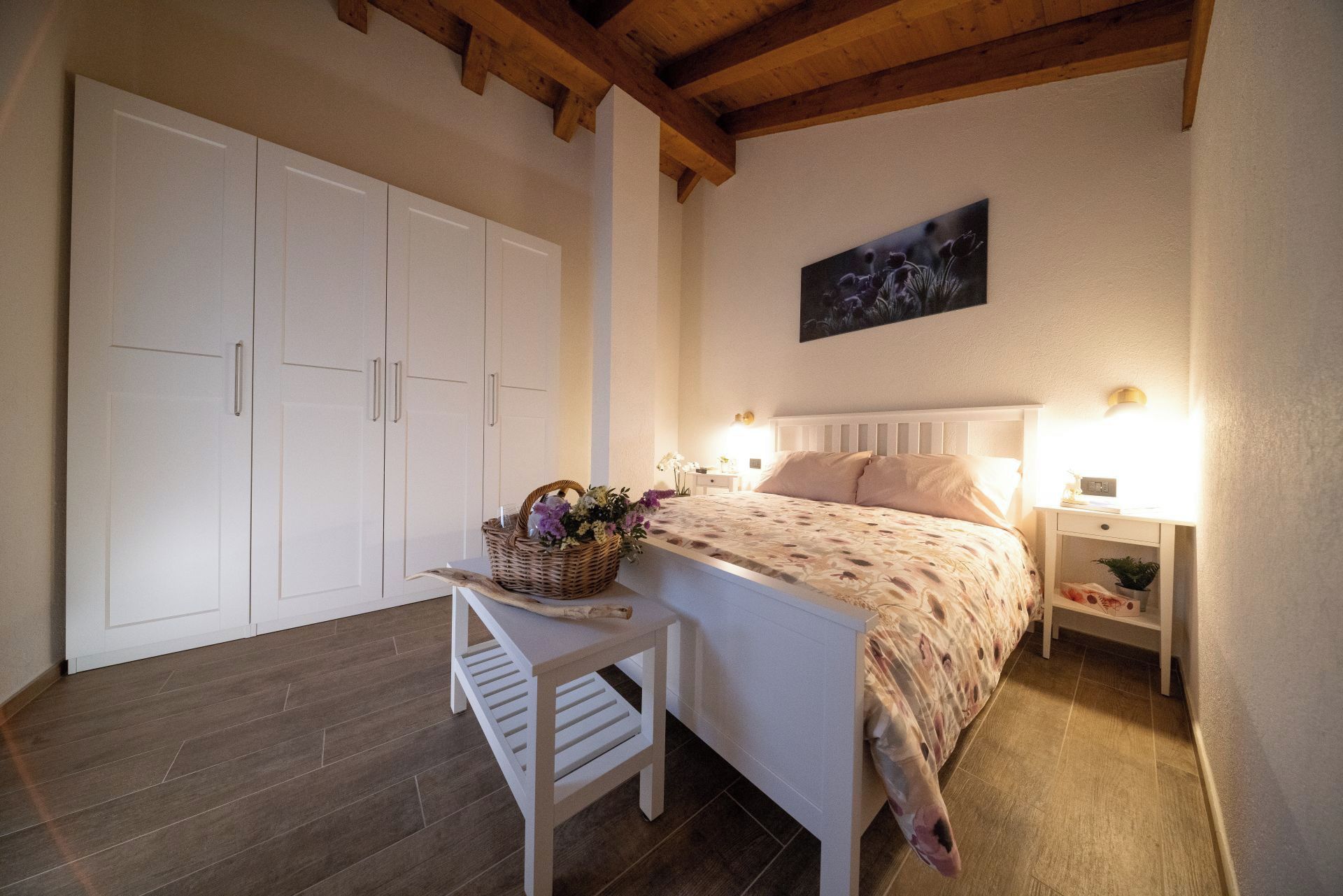 Ein Schlafzimmer mit einem Bett, Nachttischen, einem Schrank und einem Stuhl. Die Decke besteht aus Holzbalken.