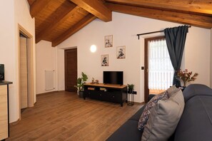 Un salon avec un plafond en bois, un canapé noir, un téléviseur posé sur un meuble en bois et un radiateur.