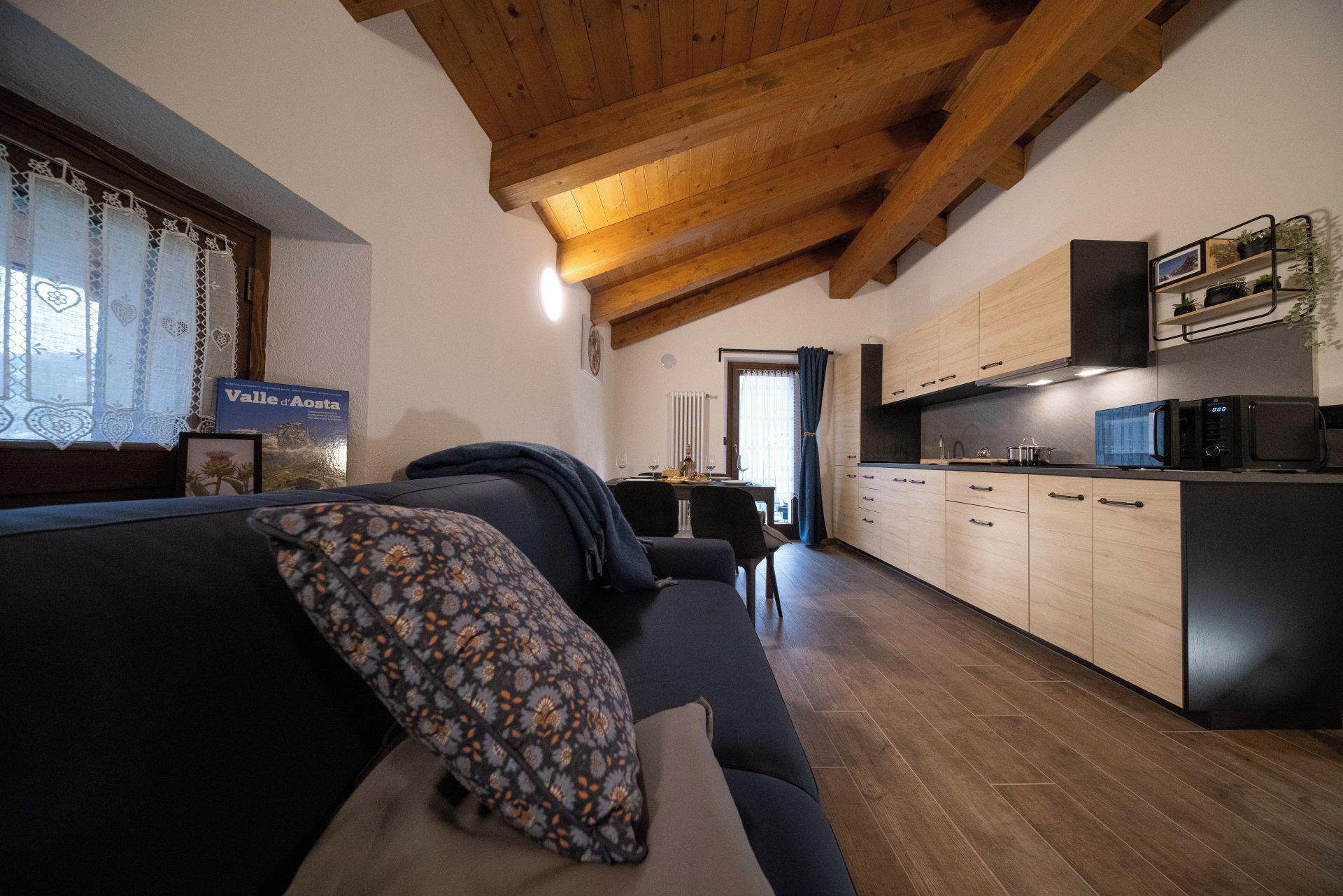 Un salon moderne avec un canapé foncé et un coussin à motifs, un coin repas avec une table et des chaises, et une cuisine avec des meubles en bois et des appareils électroménagers.