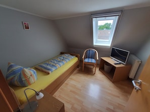 Une petite pièce, de taille modeste, comprenant un lit simple, une chaise, un bureau avec un ordinateur et une fenêtre laissant entrer la lumière naturelle.