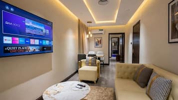 A hotel room with a flat-screen TV displaying a streaming service, a sofa, a round coffee table, and a hallway leading to a dining area.