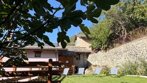 Una casa de pedra amb teulada inclinada, una tanca de fusta i un jardí amb gespa i seients de relaxació.