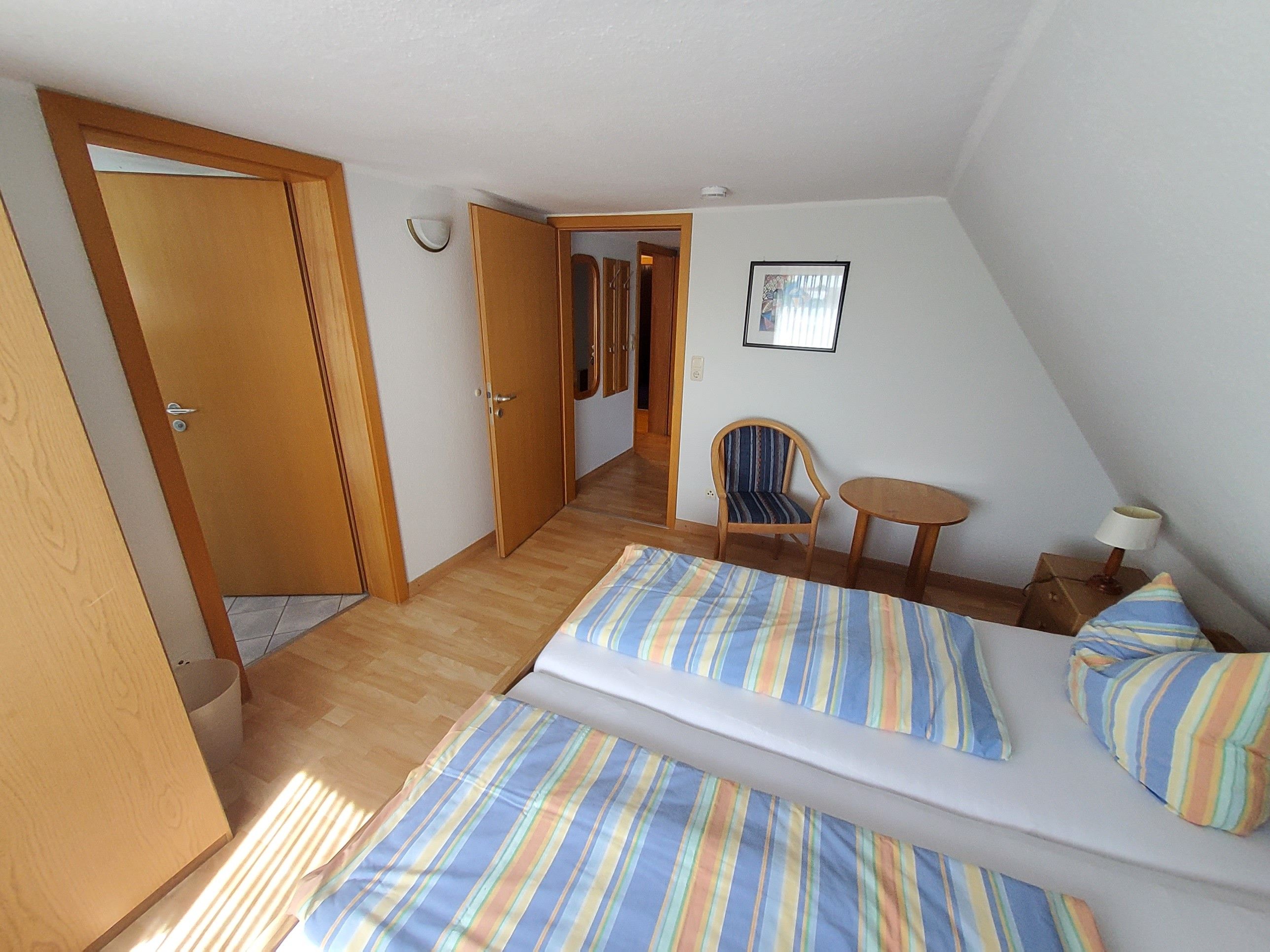 Ein Zimmer mit zwei Betten, einem Sessel, einem kleinen Tisch und einer Tür zu einem weiteren Raum.