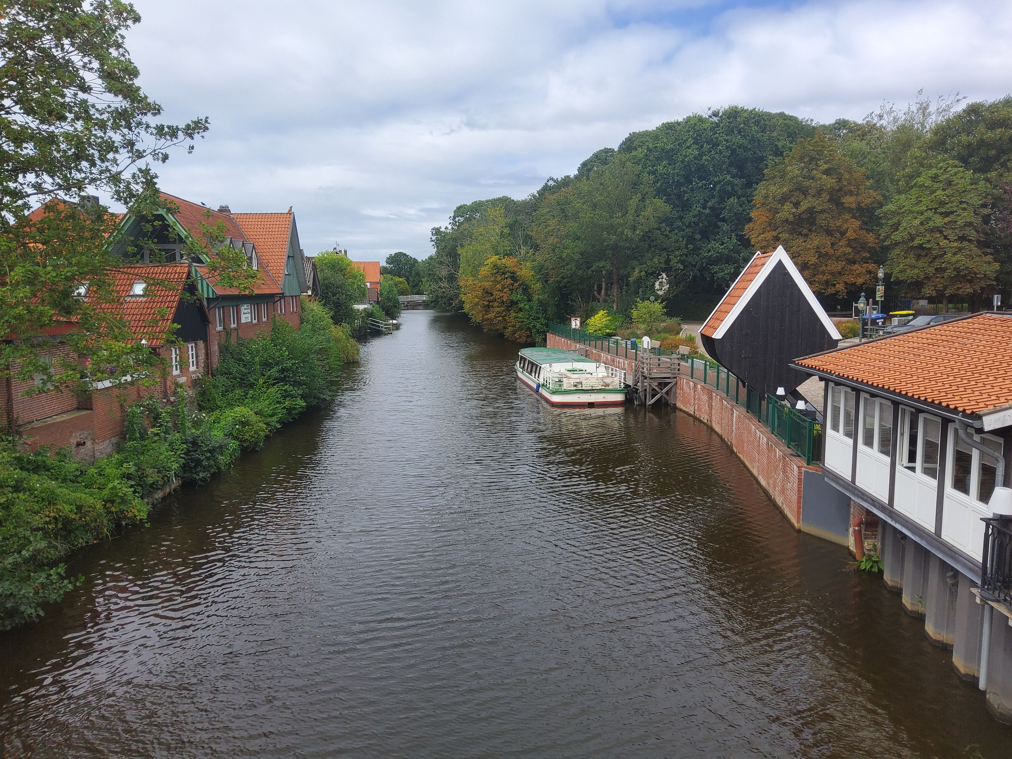 A narrow waterway flanked by buildings with red-tiled roofs, a boat docked on the right, and a bridge in the distance.