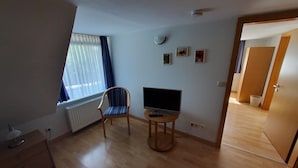 Een kamer met een houten vloer, een stoel, een televisie op een klein tafeltje, een radiator en een raam met gordijnen.