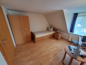 Ein Schlafzimmer mit einem Bett, einem Nachttisch, einem Schreibtisch mit Computer und einem Fenster mit Vorhängen.