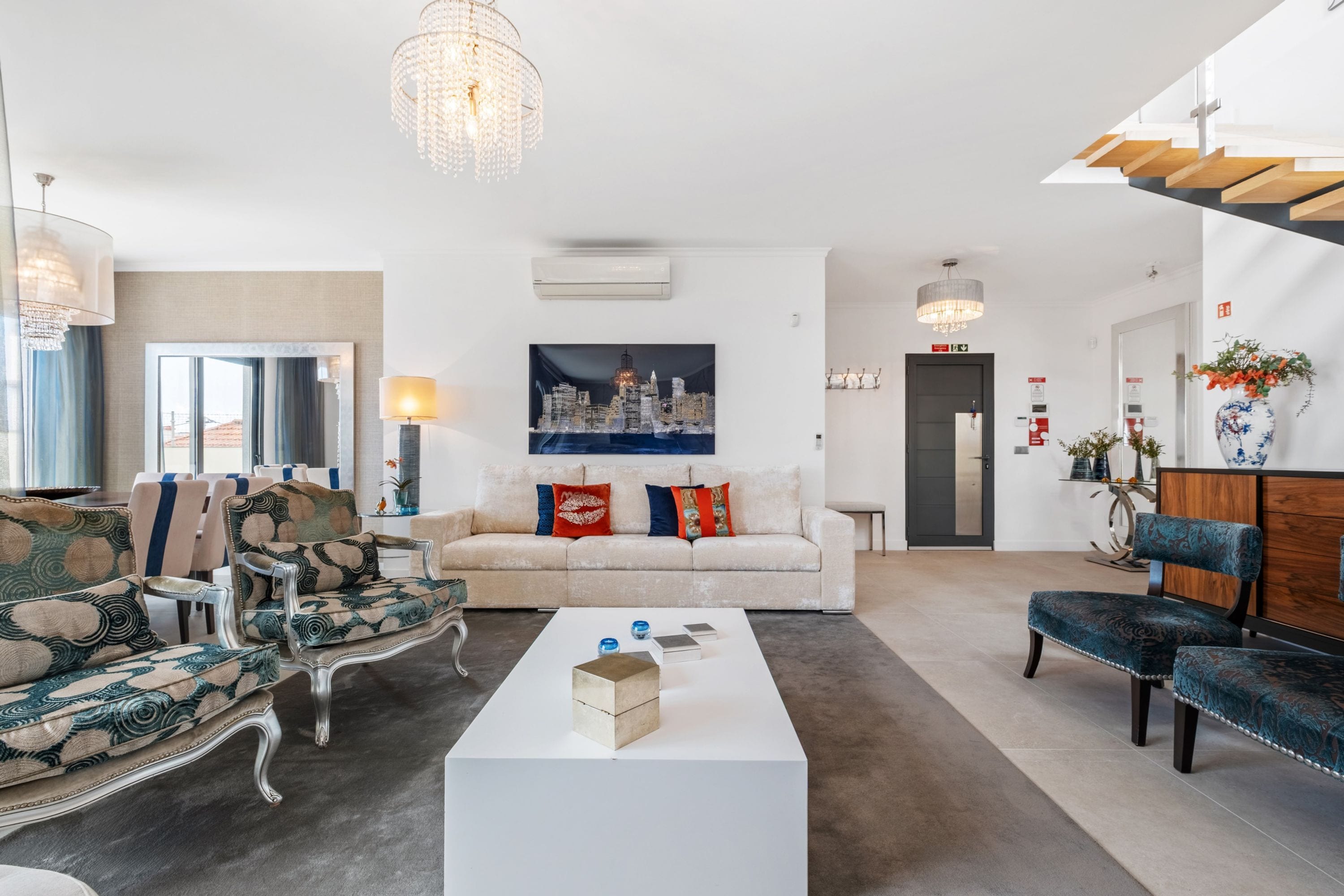 Un salon moderne avec un canapé blanc, des fauteuils à motifs, une table basse et un tableau représentant un paysage urbain.