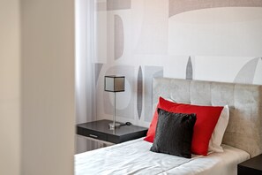 Ein ordentlich bezogenes Bett mit einem weiß-roten Kopfkissen, eine moderne Lampe auf einem Nachttisch und eine Wand mit abstrakten Mustern.