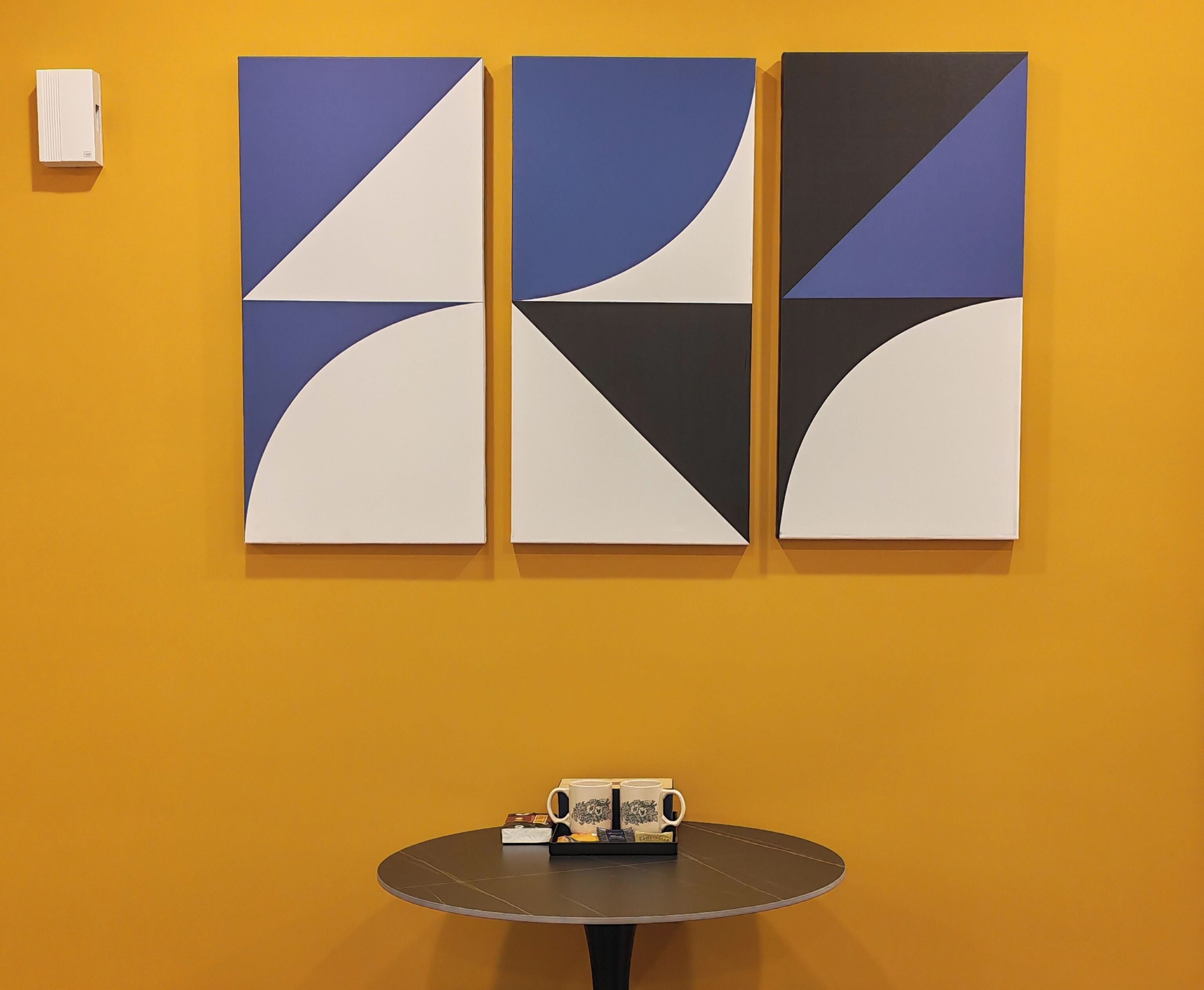 A room with a yellow wall, a round table with a tray of mugs, and three abstract paintings.