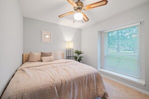 Quarto com cama, ventoinha no teto, abajur, janela com persianas e um quadro na parede.