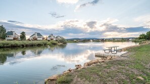 A calm river with a picnic table and a dock, surrounded by houses and a clear sky.