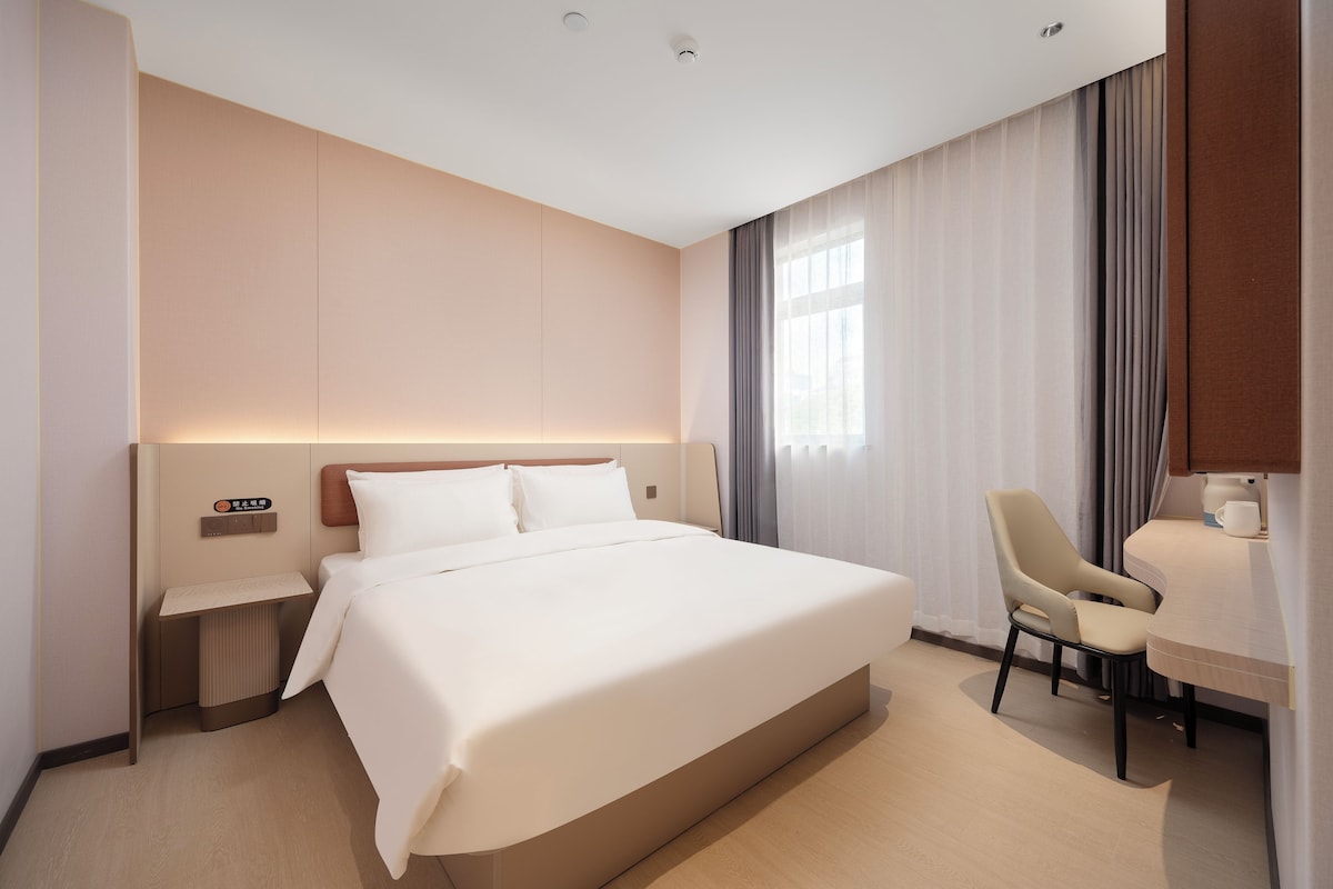 A hotel room with a bed, a desk, a chair, a nightstand, and a window with curtains.
