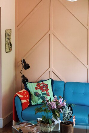 Ein Zimmer mit einem blauen Sofa, einem grünen Kissen mit roten Blumen, einer Vase mit rosa Blumen und einer Wand mit geometrischem Muster.