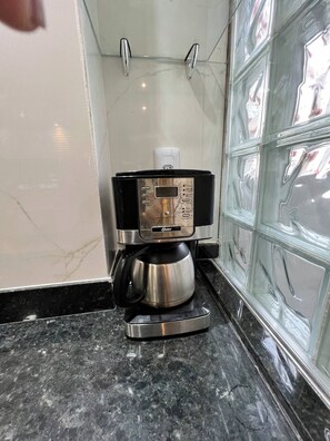 A compact coffee machine with a digital display, positioned on a black marble countertop in a corner of a room with white walls and a glass partition. - 102 Welcoming and cozy (Belém)