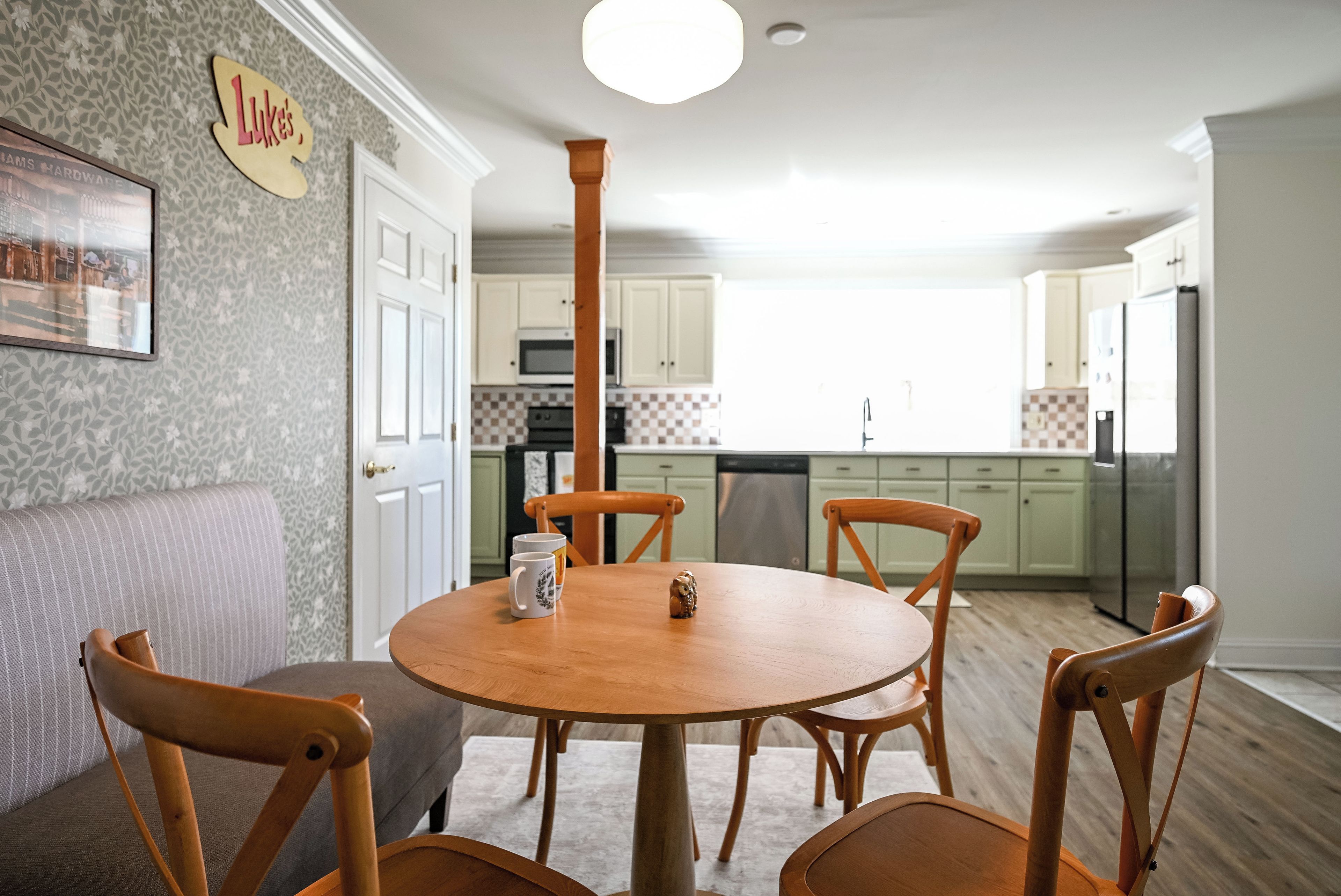 A dining area with a round wooden table, four chairs, and a sofa. A kitchen with white cabinets and a microwave is visible in the background.
