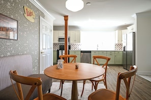 A dining area with a round wooden table, four chairs, and a sofa. A kitchen with white cabinets and a microwave is visible in the background.