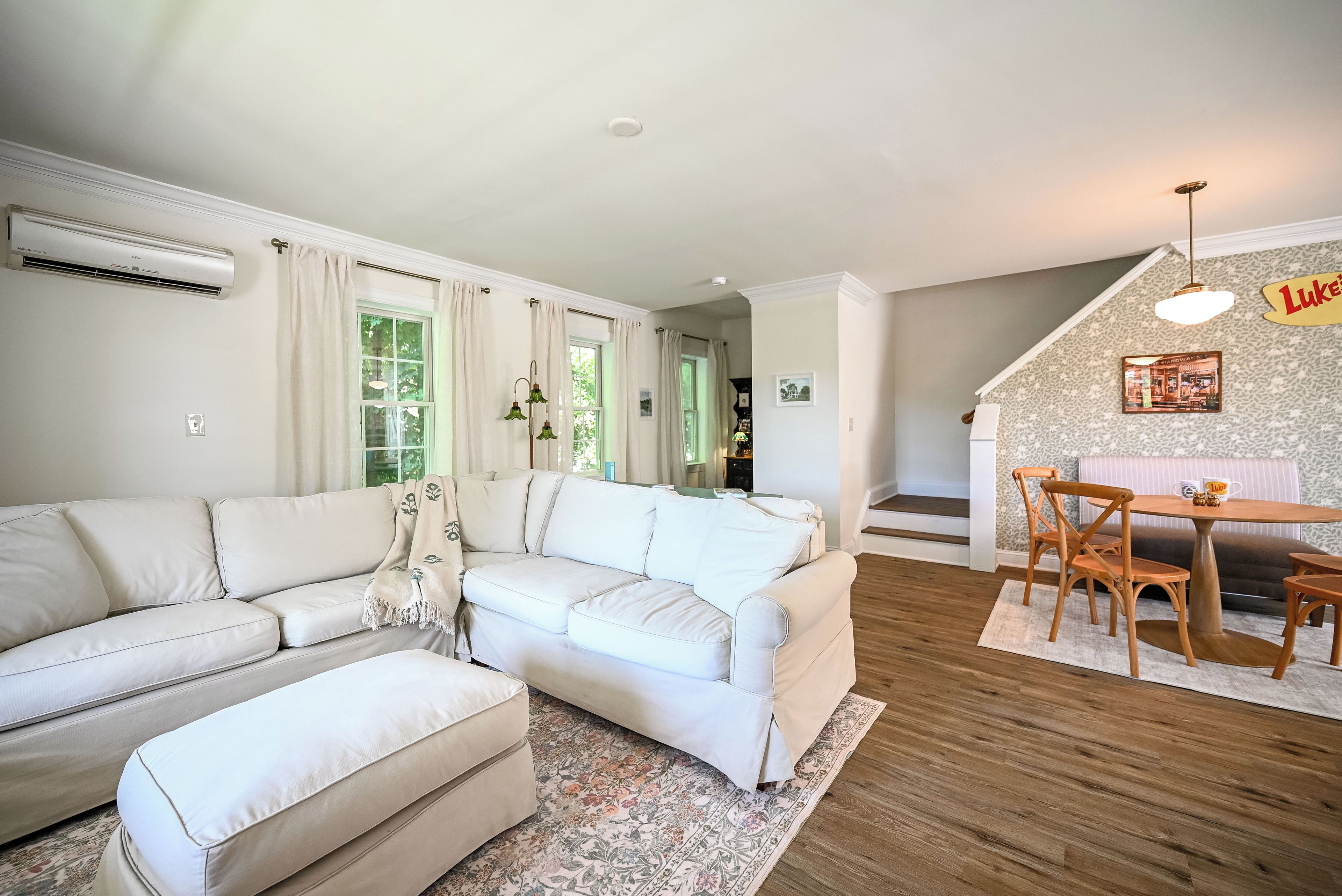A living room with a white sectional sofa, a dining area with a wooden table and chairs, and a staircase leading to an upper level.