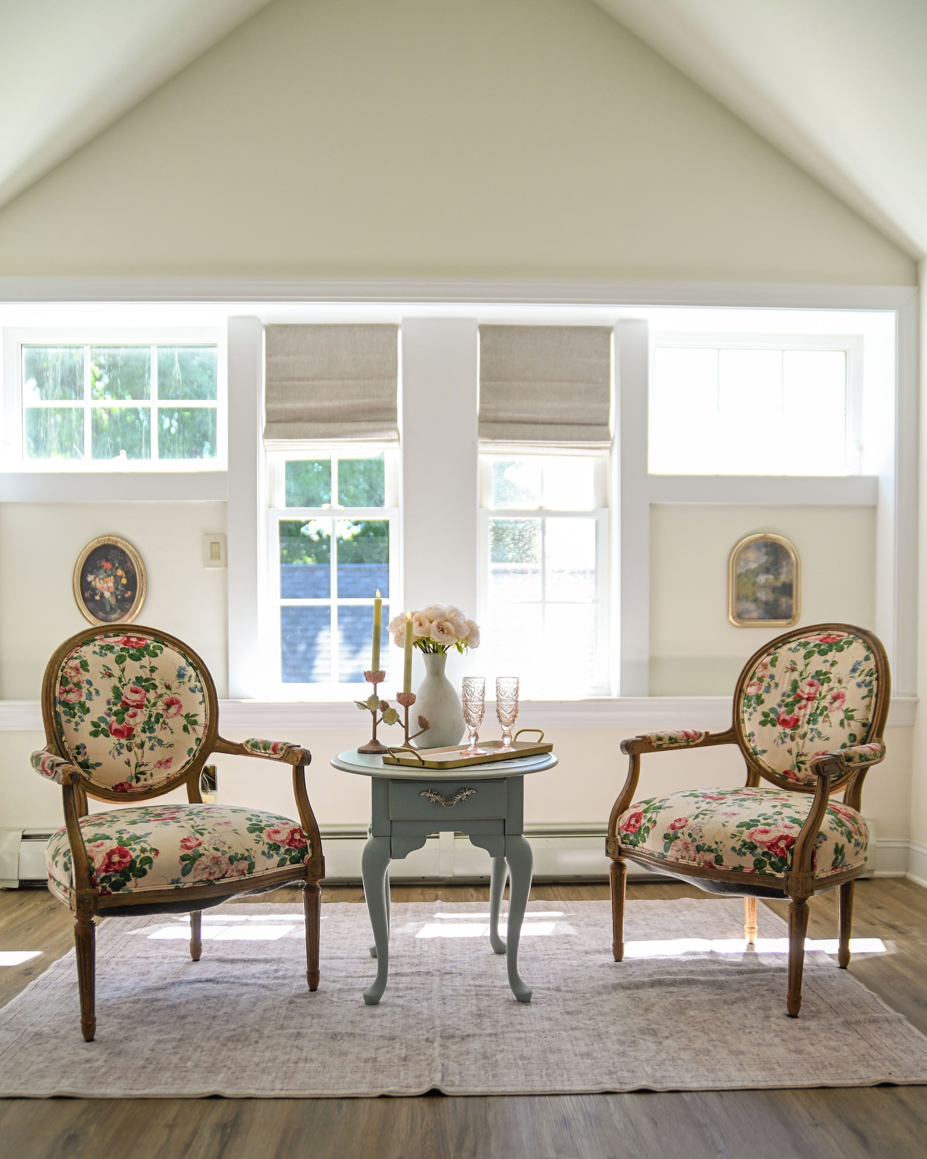 Une pièce comprenant deux fauteuils recouverts d’un tissu à motifs floraux, une petite table ornée d’un vase et de bougies, et offrant une vue dégagée grâce à de grandes fenêtres.