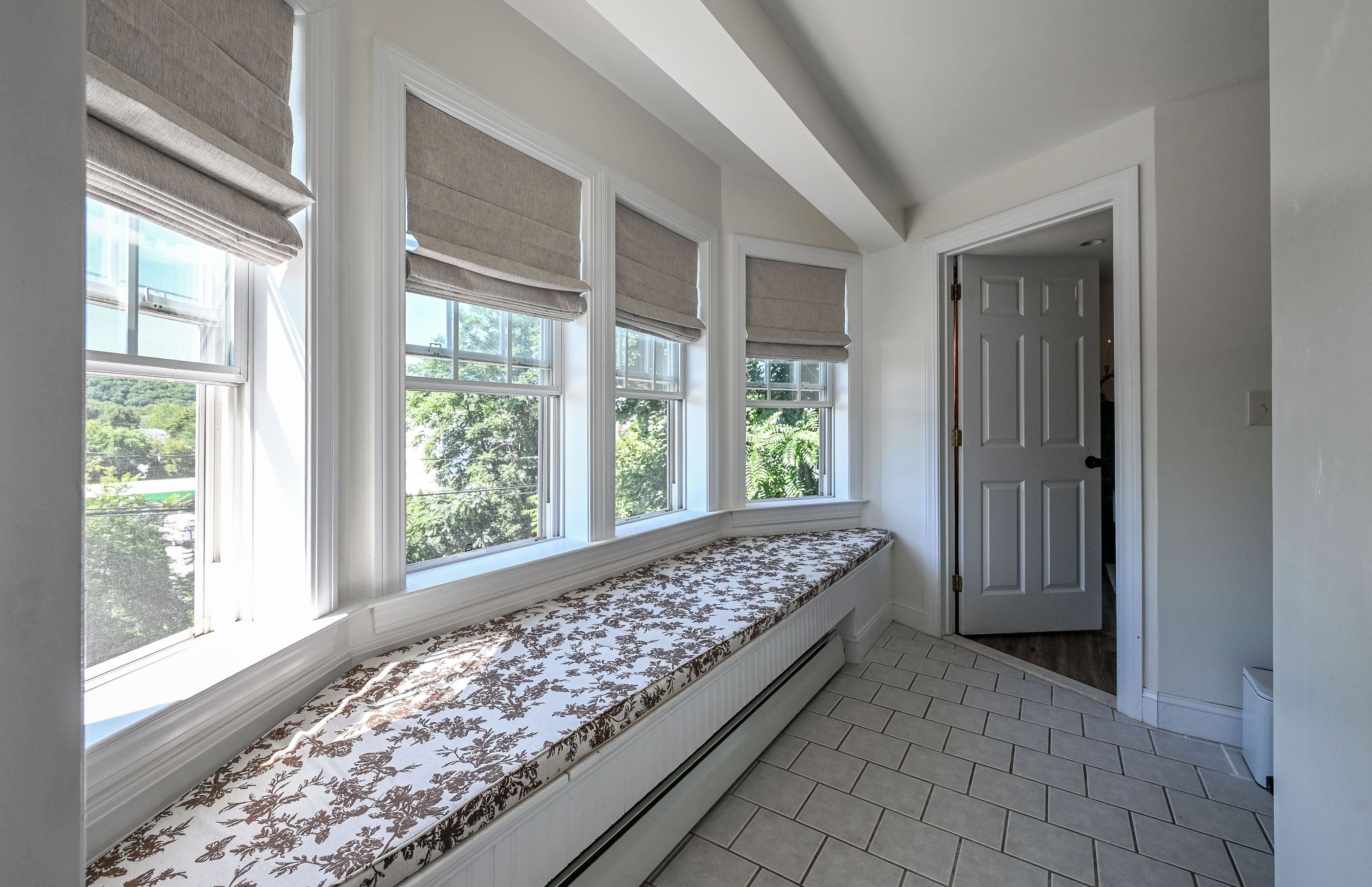 A room with a built-in bench along a wall with large windows, a door, and a tiled floor.