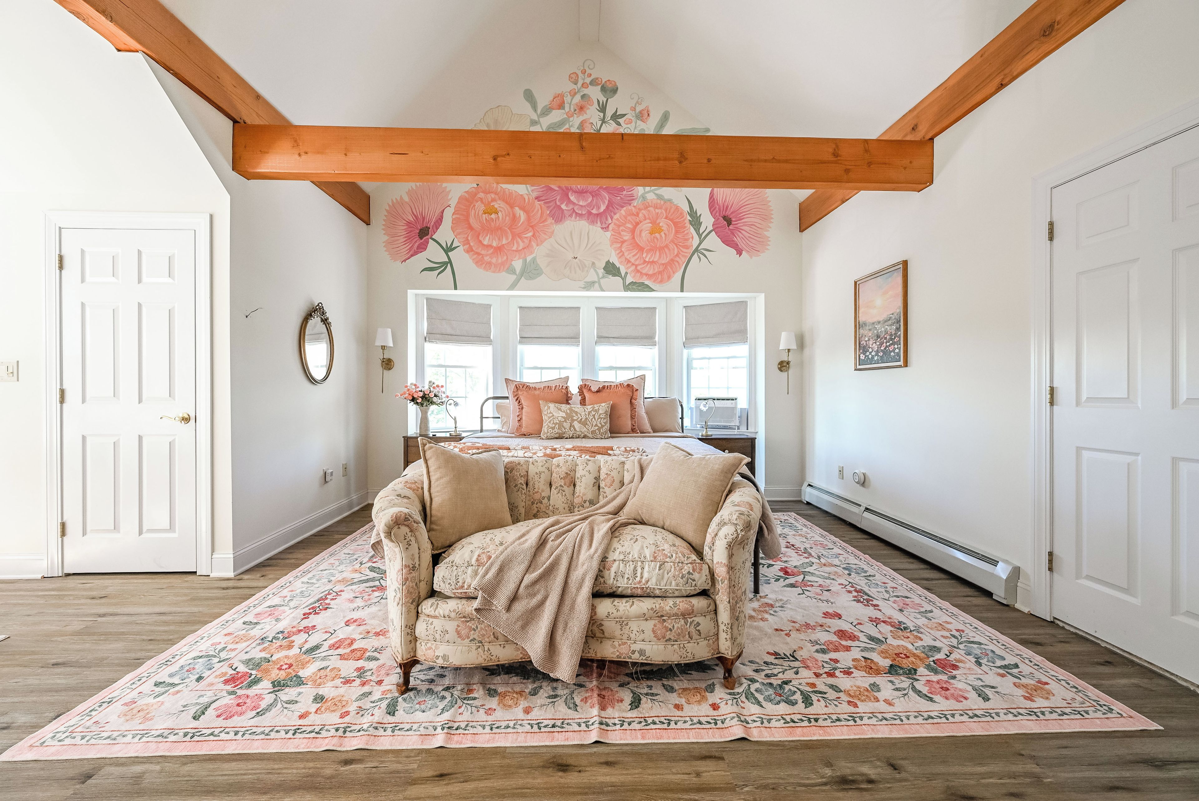 Una habitación con un sofá estampado con motivos florales, un ventanal con persianas, una viga de techo de madera y un mural floral en la pared.