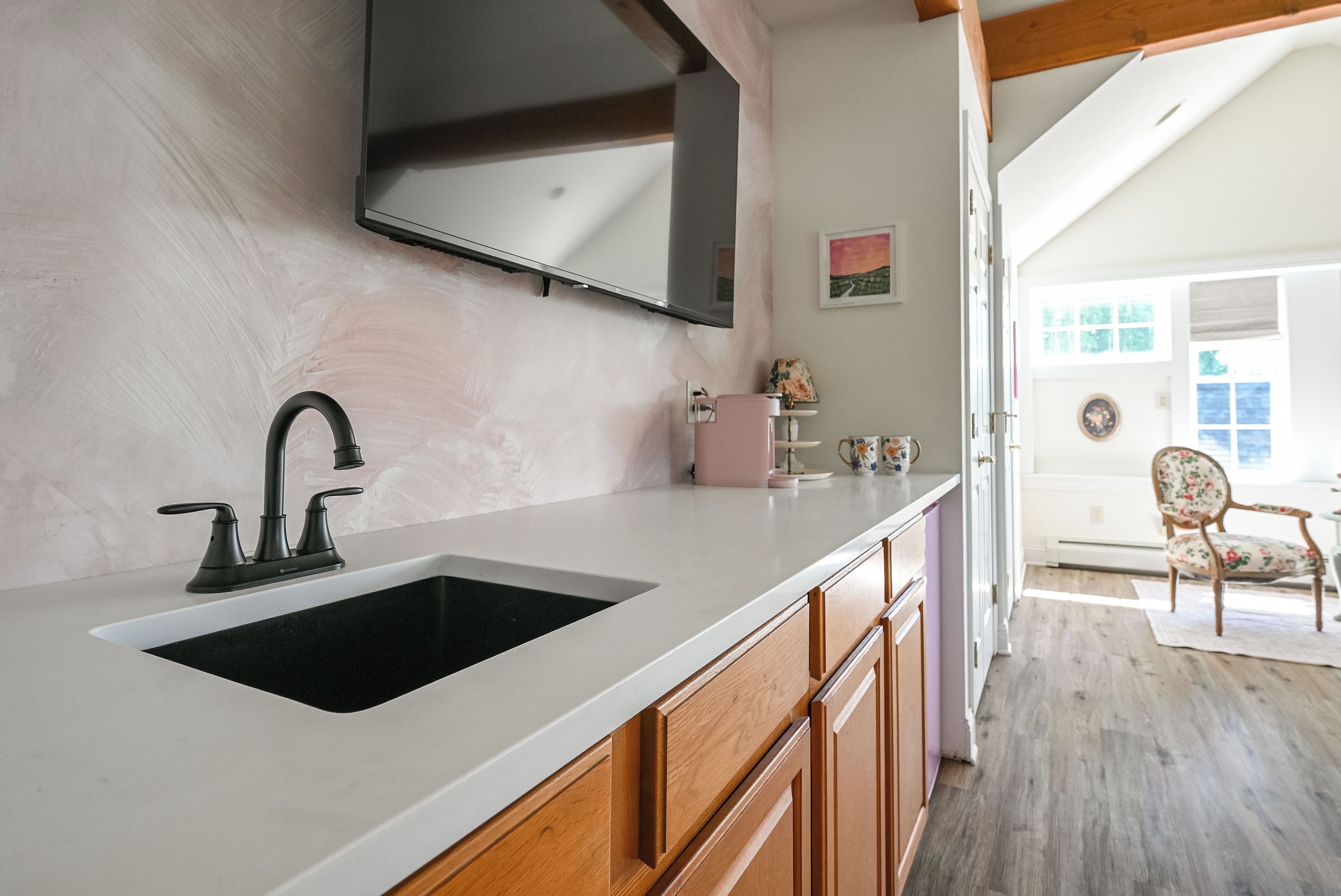 A kitchen with a white countertop, a black sink, wooden cabinets, and a mounted television.