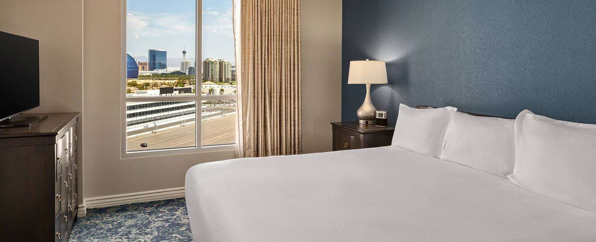 Una habitación de hotel con una cama grande, una mesita de noche con lámpara, un aparador y una ventana con vistas a la ciudad.
