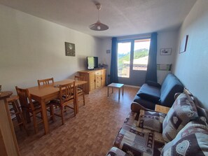 Uma sala de estar com mesa de jantar de madeira, cadeiras, sofá, televisão e vista para uma paisagem montanhosa por uma grande janela.